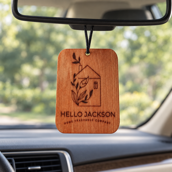 A rectangular wooden car air freshener hangs from the rearview mirror inside a car. The wood tag is engraved with a house and leaves and the text 'HELLO JACKSON HOME FRAGRANCE COMPANY'.